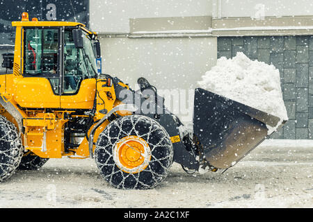 Big loader Maschine mit Stahl Ketten aus Metall entfernen von großen Schnee Haufen vom city street bei alpine Bergwelt im Winter. Starker Schneefall Nachwirkungen Stockfoto