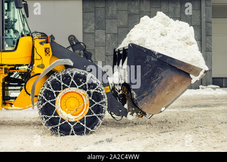 Big loader Maschine mit Stahl Ketten aus Metall entfernen von großen Schnee Haufen vom city street bei alpine Bergwelt im Winter. Starker Schneefall Nachwirkungen Stockfoto