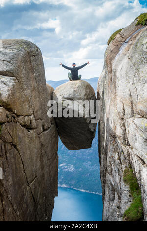 Kerl sitzt auf dem berühmten Felsen Kjerag Lysebotn Golf mit Händen raisedlooking an Fjorden und den See weiter unten. Wunderschönes Wetter mit blauem Himmel Stockfoto