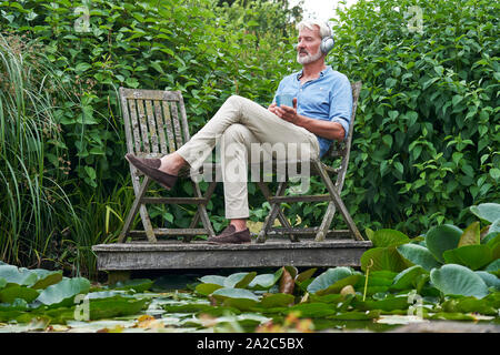 Reifer Mann Entspannen im Garten hören die Musik auf die drahtlosen Kopfhörer am Steg am See Stockfoto