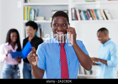 Jubelnde afrikanische amerikanische Geschäftsmann mit Bindeband am Telefon mit Kollegen im Büro der Firma Stockfoto