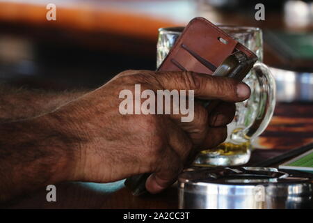 In der Nähe der Hände des Menschen mittels Smart Phone in Ledertasche mit teilweise Glas Bier getrunken und Edelstahl Aschenbecher auf Holz Balken oben Stockfoto