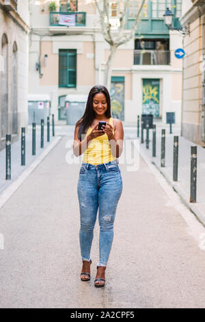 Latin touristische Mädchen mit Travel Koffer ist zu Fuß auf die Straße und über ein Mobiltelefon mit einem Hotel zu buchen. Stockfoto