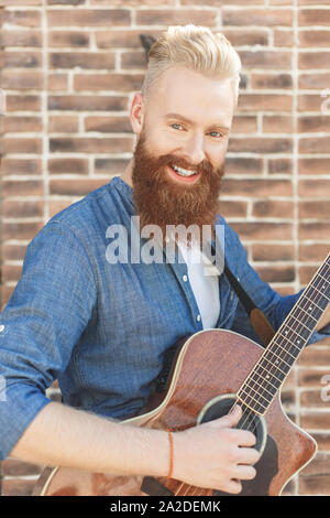 Portrait des Musikers. Hübscher junger bärtiger Mann mit akustischer Gitarre über Ziegel Hintergrund Stockfoto
