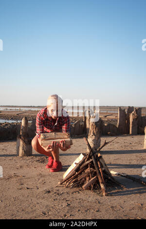 Reife blonde Frau mittleren Alters sammelt Brennholz für ein Lagerfeuer am Strand, selektiver Fokus Stockfoto