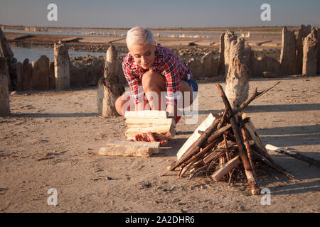 Reife blonde Frau mittleren Alters sammelt Brennholz für ein Lagerfeuer am Strand, selektiver Fokus Stockfoto