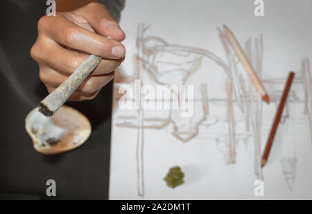 Eine gemeinsame Rauchen von Marihuana während einer Pause nach Zeichnung. Nahaufnahme von Händen und gemeinsame, blur Effekt. Hintergrund mit Bleistifte und Skizzen, Copyspace. Stockfoto