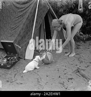 Werbefoto für Klepper Zelt: Getreu dem Werbeslogan Fahre fröhlich in die weite Welt, mit Klepper-Mantel,-Boot-und-Zelt spielt eine junge Frau mit einem Welpen vor einem Zelt, Deutschland 1930er Jahre. Werbung für ein Klepper Zelt: Eine junge Frau spielt mit einem Hund vor einem Zelt, Deutschland 1930. Stockfoto