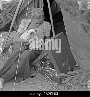 Werbefoto für Klepper Zelt: Getreu dem Werbeslogan Fahre fröhlich in die weite Welt, mit Klepper-Mantel,-Boot-und-Zelt sitzt eine junge Frau mit einem Welpen vor einem Zelt, Deutschland 1930er Jahre. Werbung für ein Klepper Zelt: Eine junge Frau sitzt mit einem Welpen vor einem Zelt, Deutschland 1930. Stockfoto