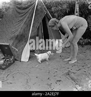 Werbefoto für Klepper Zelt: Getreu dem Werbeslogan Fahre fröhlich in die weite Welt, mit Klepper-Mantel,-Boot-und-Zelt spielt eine junge Frau mit einem Welpen vor einem Zelt, Deutschland 1930er Jahre. Werbung für ein Klepper Zelt: Eine junge Frau spielt mit einem Hund vor einem Zelt, Deutschland 1930. Stockfoto