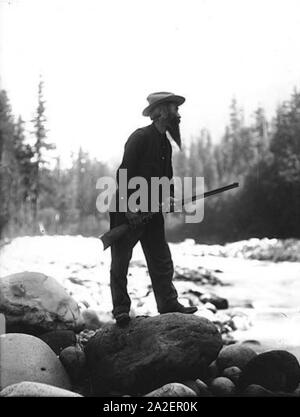 Elcaine Longmire mit Gewehr stehend auf große Boulder in Riverbed Mount Rainier National Park Washington ca 1907 (BAR30). Stockfoto