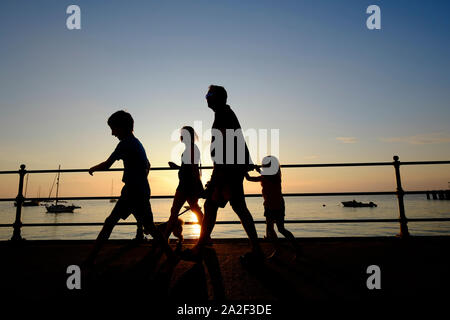 Eine Familie in Silhouette im Urlaub Wandern entlang der Verkleidung Promenade mit ihren Hund bei Sonnenuntergang in Totland Bay auf der Isle of Wight Stockfoto