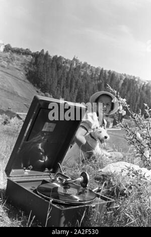 Eine junge Frau hört sich eine Platte der Schallplattenfirma Electrola mit dem Koffergrammophon auf der Wiese ein, Deutschland 1930er Jahre. Eine junge Frau, die Wiedergabe einer Aufzeichnung der Plattenfirma Electrola auf einem tragbaren Wind spielte, Gramophone, Deutschland 1930. Stockfoto