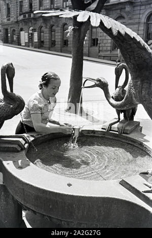 Eine junge Frau probiert das warme Thermalwasser aus dem Reiherbrunnen in der Sophienstraße der Innenstadt von Baden-Baden, Deutschland 1930er Jahre. Eine junge Frau schmeckt das warme Thermalwasser aus der Reiherbrunnen in der Sophienstraße in der Innenstadt von Baden-Baden, Deutschland 1930. Stockfoto