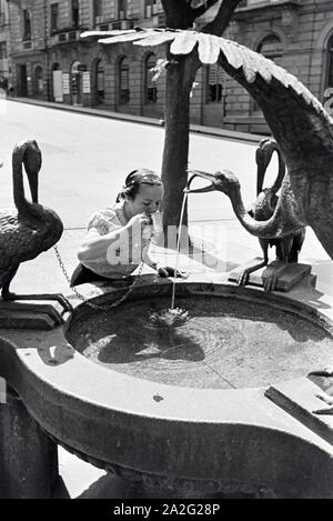 Eine junge Frau probiert das warme Thermalwasser aus dem Reiherbrunnen in der Sophienstraße der Innenstadt von Baden-Baden, Deutschland 1930er Jahre. Eine junge Frau schmeckt das warme Thermalwasser aus der Reiherbrunnen in der Sophienstraße in der Innenstadt von Baden-Baden, Deutschland 1930. Stockfoto