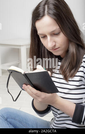 Schöne junge Schülerin Notizen mit Bleistift notebook konzentriert, sitzend auf weißem Holzboden Stockfoto