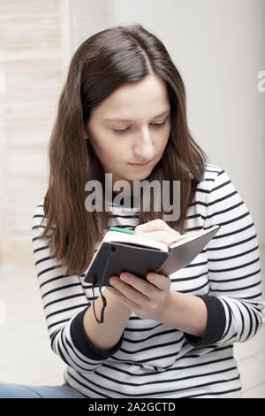 Schöne junge Schülerin Notizen mit Bleistift notebook konzentriert, sitzend auf weißem Holzboden Stockfoto