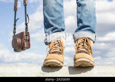 Junge Frau mit Füßen gekleidet in Stiefel, stehend auf einer Betonwand, die eine Kamera in den Händen. Vor dem Hintergrund der blauen Himmel und weiße Clou Stockfoto