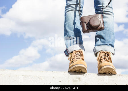 Junge Frau mit Füßen gekleidet in Stiefel, stehend auf einer Betonwand, die eine Kamera in den Händen. Vor dem Hintergrund der blauen Himmel und weiße Clou Stockfoto