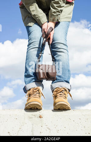 Junge Frau mit Füßen gekleidet in Stiefel, stehend auf einer Betonwand, die eine Kamera in den Händen. Vor dem Hintergrund der blauen Himmel und weiße Clou Stockfoto