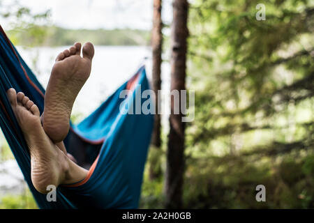 Die Füße des Menschen sind in der Nähe gesehen - aus einer Hängematte im Sommer Wald, auf einem unscharfen Hintergrund aus Pinien und einem See. Stockfoto