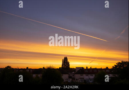 Wimbledon, London, UK. 3. Oktober 2019. Gelbe Sonnenaufgang beleuchtet ein hoher Höhe Vapor Trail gegen den blauen Himmel über Suburban Dächer in South West London. Credit: Malcolm Park/Alamy Leben Nachrichten. Stockfoto