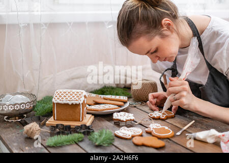 Vereisung der Weihnachtsbäckerei. Frau dekorieren Honig-konditorenlebkuchen Cookies auf Holz braun Tabelle. closeup, kopieren. Leere keks Gingerbread House Stockfoto