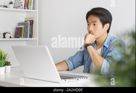 Portrait asiatischen Casual Geschäftsmann in Denim oder Jeans Shirt mit Laptop und Denken und schweren Pose im Home Office. Casual Geschäftsmann arbeiten mit tec Stockfoto