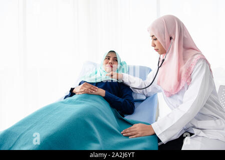 Eine junge Schöne muslimischen Arzt ist mit einem Stethoskop auf die Herzfrequenz des Patienten am Bett im Krankenhaus Untersuchungsraum zu hören. Stockfoto