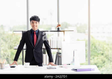 Porträt einer zuversichtlich glückliche junge Geschäftsmann im Anzug, der an einem Tisch in einem modernen Büro. Stockfoto