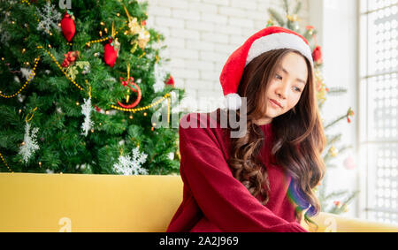 Eine Frau trägt ein Santa Hut in einem roten Kleid mit einem Weihnachtsbaum auf der Rückseite. Das schöne Mädchen lächelt und wartet auf das Weihnachtsgeschenk von h Stockfoto