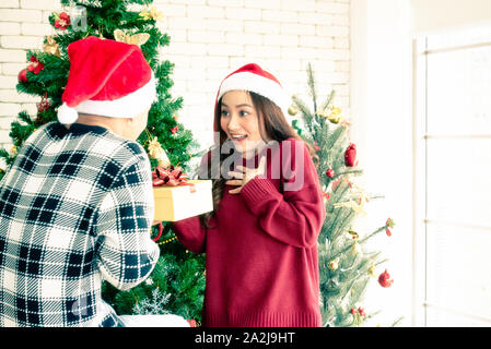 Ein paar mit Santa's Hut im Zimmer, eine schöne Frau ist glücklich und überrascht, als sie ein gelbes Geschenk Box von Ihrem Freund zu Weihnachten bekommen. Co Stockfoto