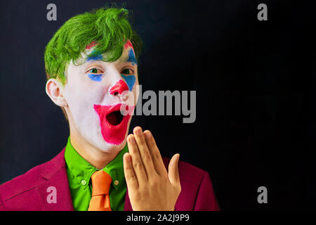 Mann in Mime makeup Cosplay mit grünen Haaren und einem roten Anzug eine orangefarbene Krawatte und ein grünes T-Shirt. Clown überrascht oder schockiert Stockfoto