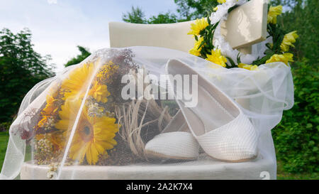 Strauß gelber Gerbera Daisy und weiße Schuhe der Braut mit einem Schleier auf einem weißen Holzstuhl im Hinterhof abgedeckt Stockfoto