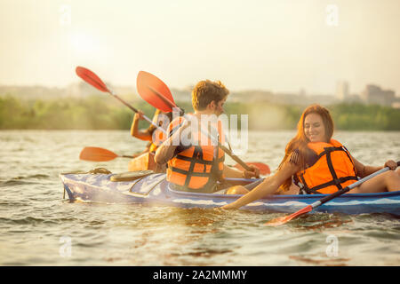 Glückliche Junge kaukasier Gruppe von Freunden Kajakfahren auf dem Fluss mit Sonnenuntergang im Hintergrund. Spaß in der Freizeit. Gerne männliche und weibliche Modell laughting auf dem Kajak. Sport, Beziehungen Konzept. Stockfoto