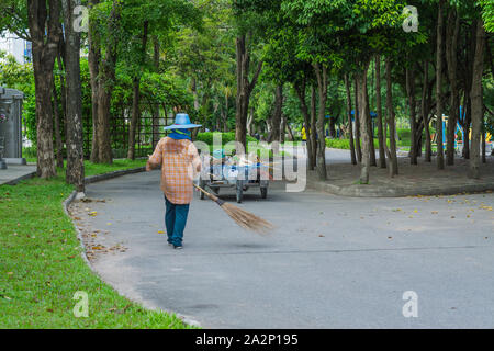 Eine asiatische Frau fegt Laub von der Straße in den Garten. Reinigung Konzept. Stockfoto