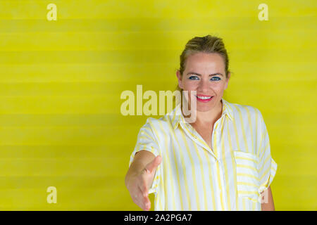 Junge schöne Frau mit gestreiften Hemd stehend über isolierte gelben Hintergrund lächelt freundlich mit Handshake als Gruß und einladend. Stockfoto