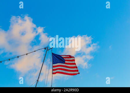 Eine amerikanische Fahne weht im Wind wie es hängt von einem gaff am Heck eines Bootes. Die Fahne fliegt vor einem blauen Himmel mit weißen Wolken. Stockfoto