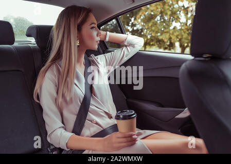 Schöne Geschäftsfrau Fahrten im Taxi im Auto, hält Schale Kaffee, Tee, Snacks Frühstück, geht die Arbeit ein wichtiges Meeting, hat Rest blickt Fenster Stockfoto