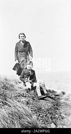 Negative - Cowes, Phillip Island, Victoria, 1938, eine Frau mit einem kleinen Jungen, der auf den Felsen am Wasser sitzt. Er hält ein Pinguin Stockfoto