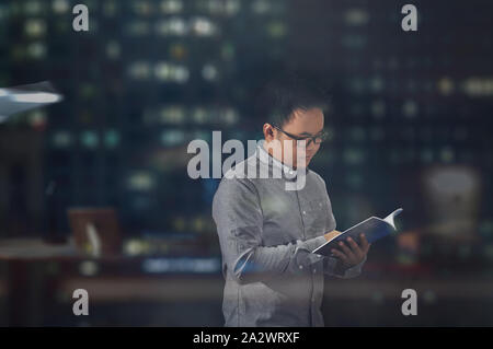 Seitenansicht geschossen von einem jungen Mann in langen Ärmeln und Brillen stehen und lesen Sie ein Buch in einem Bürogebäude in der Nacht. Stockfoto