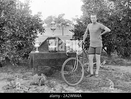 Negative - Wentworth District, New South Wales, ca. 1920, ein Radfahrer mit seinem Fahrrad und seine Trophäen, die auf einem drapiert Tabelle angezeigt wurden. Es gibt Obstbäume auf beiden Seiten von ihm und Kohl in einem Gemüsegarten vor ihm Stockfoto