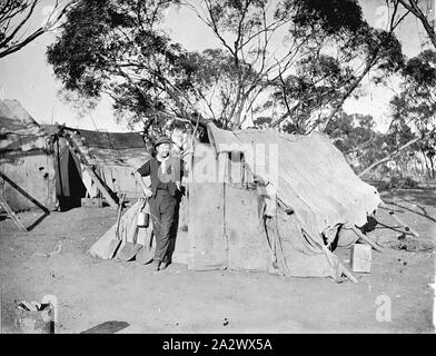 Negative - Goulburn Valley, Victoria, ca. 1915, ein Mann vor einem Zelt gebaut der Hessischen. Es gibt ähnliche Hütten hinter ihm. Ein Spaten, zwei Schaufeln und kann Billy sind hielten gegen das Zelt Stockfoto