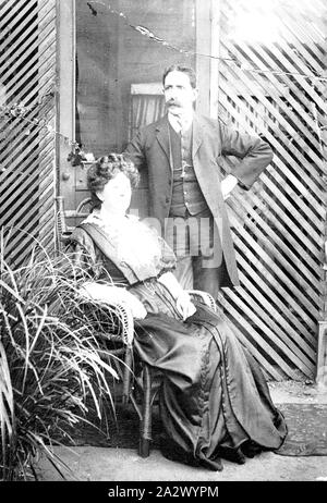 Negative - Melbourne, Victoria, ca. 1895, eine Frau in einem Zuckerrohr Stuhl sitzen, ein Mann steht neben ihr und es gibt eine Tür, die in einen Bereich geschlossen mit einem Gitter hinter Ihnen Stockfoto