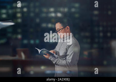 Junger Mann auf der Suche glücklich und lächelnd, während er ein Buch zu lesen. Student trägt lange Ärmel und Brillen geben positive Emotionen beim Lesen eines Buches Stockfoto