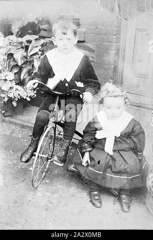 Negative - Kingower, Victoria, etwa um 1900, ein Studio Portrait von zwei kleinen Jungen. Man sitzt auf einem Dreirad, während der andere, der trägt Röcke, auf einem Stuhl sitzt. Er hält einen Penny in seiner Hand, als er noch bestochen werden zu sitzen hatte Stockfoto