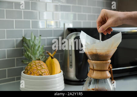 Nahaufnahme der Hand eines Mannes, der Kaffee in einer Tropfkaffeeflasche mit einer Obstschale im Hintergrund rührt. Stockfoto