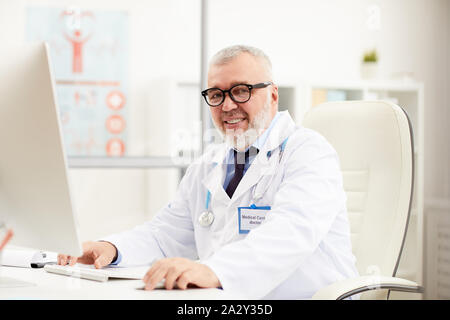 Portrait von leitenden Arzt in Brillen am Tisch sitzen und lächeln in die Kamera während der Arbeit am Computer im Büro Stockfoto