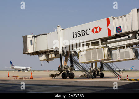 Eine Jet-Brücke mit dem HSBC-Logo auf dem belebten Flughafen Shanghai Pudong, auf dem mehrere Flugzeuge zur Abfahrt im Hintergrund auf der Startbahn stehen. Stockfoto