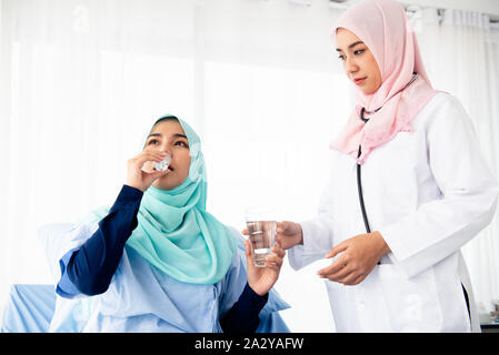 Eine schöne muslimische Frau Doktor trug ein Stethoskop, Medizin und Wasser, um den kranken Patienten auf dem Bett im Untersuchungsraum zu essen. Stockfoto